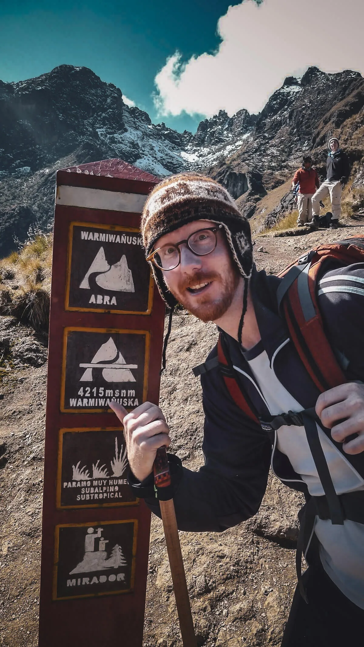 A warmly dressed hiker beside the Warmi Wañusqa pass marker with high mountains behind
